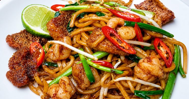 Glossy Mee Goreng Melayu with yellow noodles, tofu puffs, prawns, cabbage, and fried shallots on a banana leaf plate with lime wedge